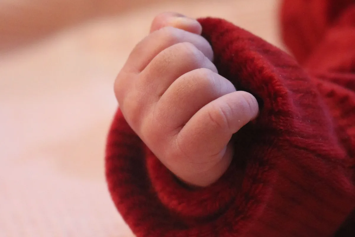 Eine kleine Babyhand schaut aus einem roten Ärmel, darunter eine helle Decke